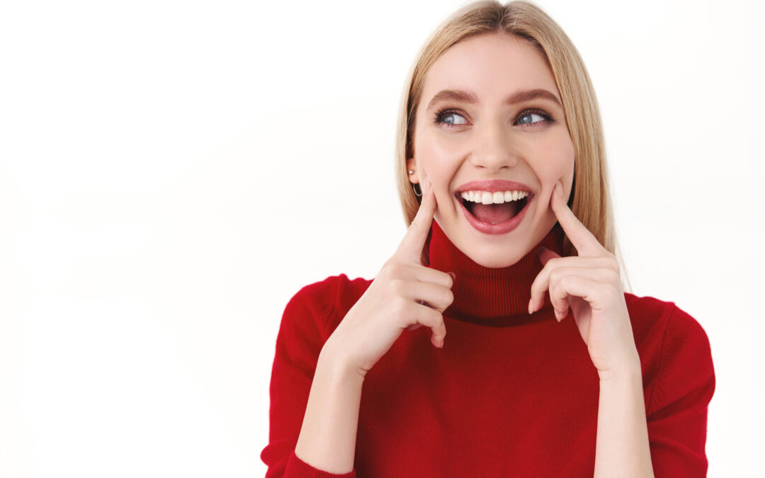 Woman with bright white smile pointing to her teeth in red turtleneck.